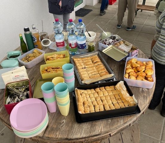 Ein Holztisch mit Lebensmitteln, einschließlich Gebäckschalen, Flaschen und Tassen, ist aufgebaut. Verschiedene Snacks und Getränke sind auf dem Tisch arrangiert. Leute stehen um den Tisch herum.