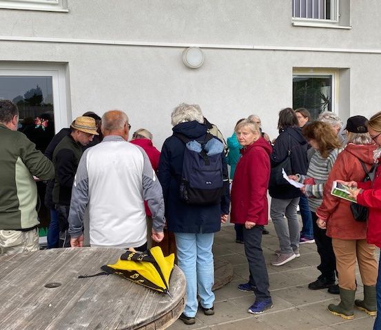 Eine Gruppe von Menschen wartet vor einem Gebäude. Einige lesen, während andere reden. Es gibt einen Holztisch mit Taschen darauf.
