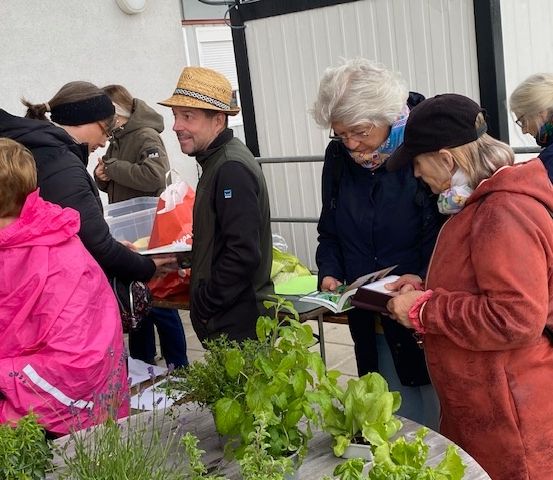 Eine Gruppe von Menschen versammelt sich um einen Tisch mit Pflanzen, und ein Mann in einem Hut spricht zu ihnen.