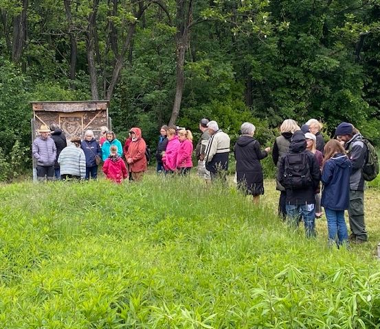 Eine Gruppe von Menschen in bunten Mänteln und Mützen versammelt sich auf einem Rasen mit einer Holzstruktur im Hintergrund.