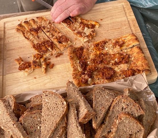 Eine Person schneidet ein Stück Pizza mit Speck auf einem Holzschneidebrett. Unten ein Korb mit Vollkornbrot.