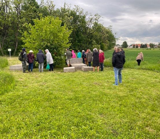 Eine Gruppe von Menschen in verschiedenen Jacken und Mützen steht auf einer Wiese, einige auf einer Holzplattform. Bäume und ein bewölkter Himmel im Hintergrund.