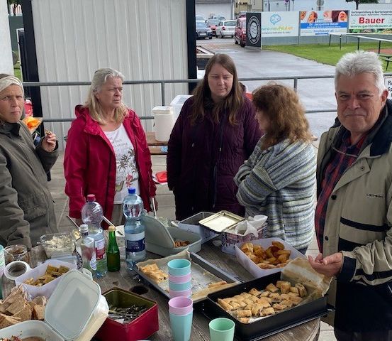 Fünf Menschen stehen um einen Tisch voller verschiedener Speisen. Der Mann auf der rechten Seite sieht ernst aus, während die Frau auf der linken Seite raucht. Im Hintergrund sind geparkte Autos und ein Gebäude mit Bannern zu sehen.