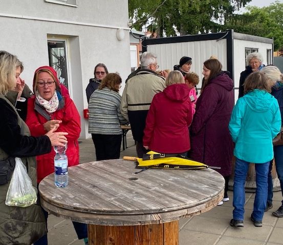 Eine Gruppe von Erwachsenen, möglicherweise bei einem Gemeindetreffen, steht um einen Holztisch mit einem gelben Schirm herum. Einige tragen Winterkleidung. Eine Frau trägt eine Plastiktüte. Bäume und ein Gebäude sind im Hintergrund zu sehen.