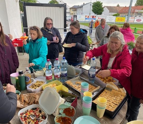 Eine Gruppe von Frauen versammelt sich um einen Tisch im Freien und genießt Essen und Getränke. Auf dem Tisch befinden sich verschiedene Gerichte, Teller, Becher und Flaschen. Eine der Frauen hält einen Löffel. Im Hintergrund stehen ein Gebäude, Bäume und ein Rasenfeld.