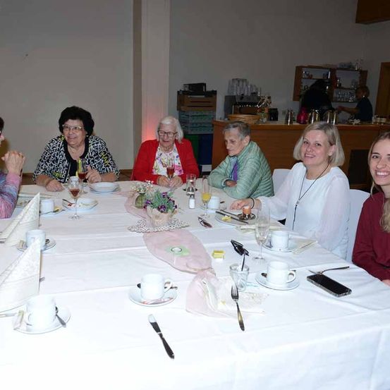 Eine Gruppe von Frauen sitzt um einen Tisch herum, lächelt und unterhält sich. Der Tisch ist mit Tellern, Weingläsern, Tassen und Besteck gedeckt. Ein rosa Tischläufer verleiht dem Ganzen eine dekorative Note.