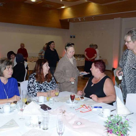 Eine Gruppe von Frauen sitzt um einen Tisch in einem Bankettsaal. Der Tisch ist mit Gläsern, Tellern und einer Blumendekoration besetzt. Eine Frau steht hinter dem Tisch und hält einen Teller. Im Hintergrund steht eine Frau vor einem Buffettisch.