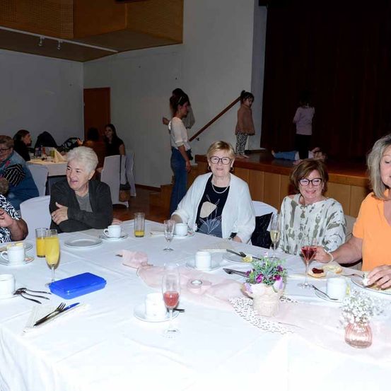 Eine Gruppe von Frauen sitzt an einem Tisch in einem Speisesaal. Sie tragen Brillen und lächeln. Der Tisch ist mit Tellern, Tassen und einem Blumenvase gedeckt. Dahinter befinden sich eine Treppe und eine Bühne. Einige Leute stehen auf der Bühne.