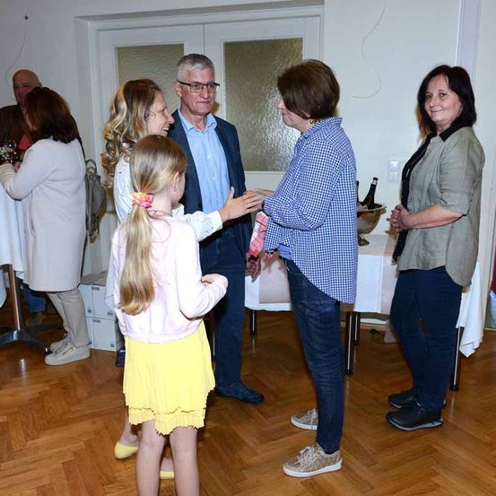 Mehrere Erwachsene und ein junges Mädchen versammeln sich in einem Raum. Ein Mann mit Brille und eine Frau mit kariertem Hemd stehen in der Mitte und unterhalten sich. In der Nähe lächelt eine Frau in einer beigen Jacke das Mädchen an. Ein Tisch mit Weinflaschen steht hinter ihnen.