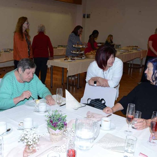 Eine Gruppe von Frauen genießt eine Mahlzeit an einem Tisch mit verschiedenen Speisen und Weingläsern. Eine Frau isst, während andere sich unterhalten. Im Hintergrund sind Tische mit Essen und anderen Leuten zu sehen.