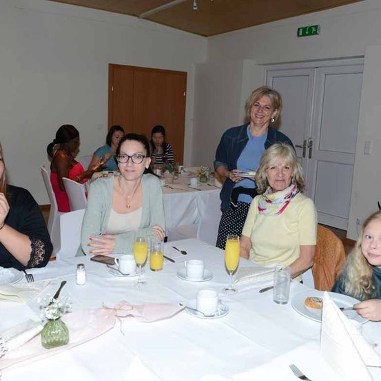 Eine Gruppe von Frauen sitzt an einem Tisch mit Getränken und Essen, lächelt und interagiert. Der Tisch ist mit einer weißen Tischdecke bedeckt, und es gibt Gläser, Tassen und Teller. Eine Frau steht in der Nähe und hält einen Teller. Der Raum hat weiße Wände und Türen.