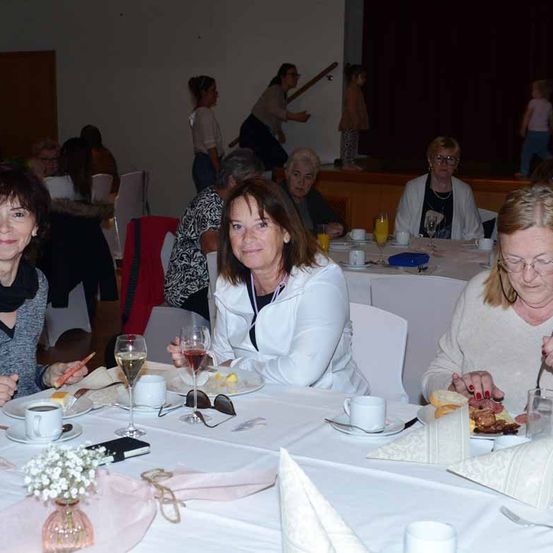 Mehrere erwachsene Frauen sitzen an einem Tisch und genießen ihr Frühstück. Eine Frau in der Mitte hält ein Weinglas, während andere essen und plaudern. Dahinter sitzen oder stehen weitere Personen.