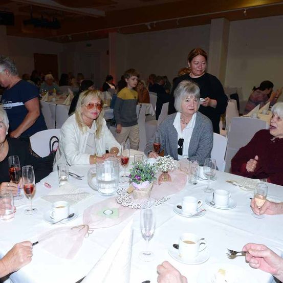 Eine Gruppe von Menschen sitzt um einen Tisch mit Weingläsern, Tassen und Blumen. Eine Frau hält einen Teller, und andere lächeln. Eine Person steht in der Nähe des Tisches, und andere unterhalten sich im Hintergrund.