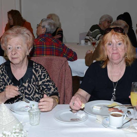 Zwei ältere Frauen sitzen an einem Esstisch. Eine isst, während die andere ein Glas Saft hält. Sie gehören zu mehreren Menschen in einem Raum mit Tischen und Stühlen.