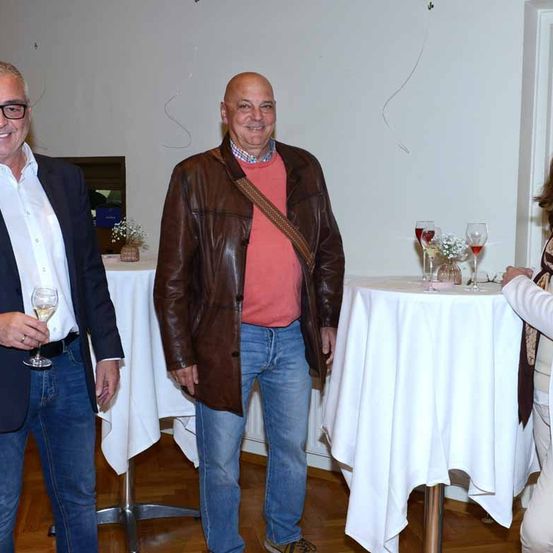 Drei Männer stehen in einem Innenraum, möglicherweise bei einer Party. Der Mann auf der linken Seite hält ein Weinglas. Der Mann in der Mitte lächelt und schaut den Mann auf der rechten Seite an. Der Tisch vor ihnen hat Weingläser und eine Blumenarrangement.