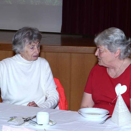 Zwei ältere Frauen sitzen an einem Tisch und unterhalten sich. Eine trägt ein weißes Rollkragenshirt, die andere ein rotes Shirt mit einem herzförmigen Dekor. Der Tisch ist mit einer Schüssel, einem Becher und Besteck gedeckt. Dahinter befindet sich eine Holzbühne mit einer Projektionsleinwand.