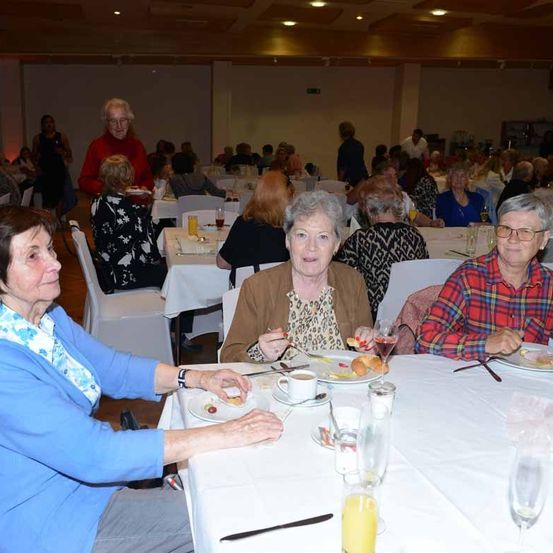 Eine Gruppe älterer Frauen sitzt an einem Esstisch, möglicherweise in einem Restaurant. Sie essen und unterhalten sich miteinander. Der Tisch ist mit weißen Tischdecken bedeckt und hat verschiedene Gegenstände darauf. Der Raum ist schwach beleuchtet.