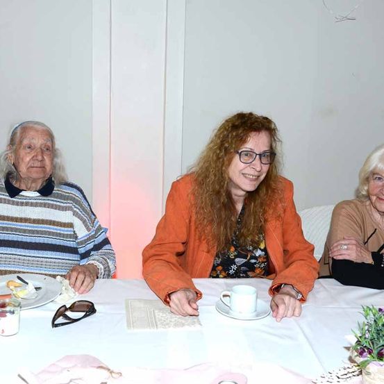 Drei ältere Frauen sitzen um einen Tisch. Die Frau in der Mitte trägt eine orangefarbene Jacke und eine Brille und lächelt. Die Frau auf der linken Seite hat einen gestreiften Pullover und einen Teller mit Essen. Die Frau auf der rechten Seite trägt einen Ring.