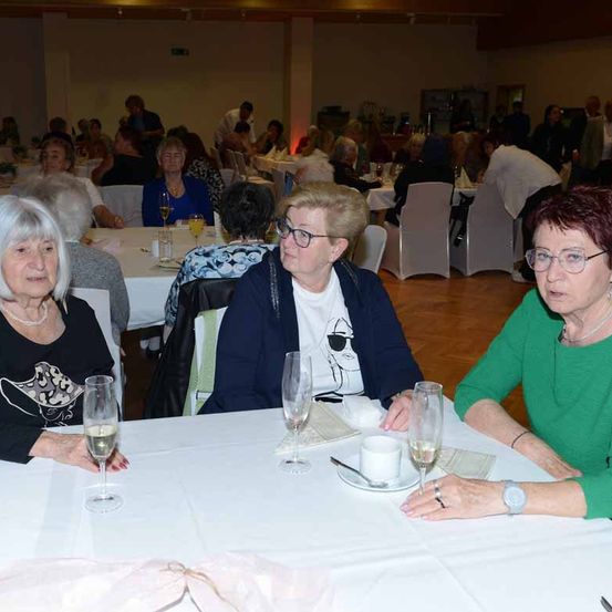 Mehrere ältere Frauen sitzen in einem Speisesaal an einem Tisch und unterhalten sich. Jede hat ein Weinglas. Im Hintergrund sitzen andere Gäste mit weißen Stühlen und Tischen.