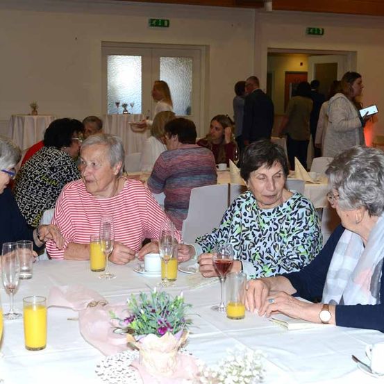 Mehrere ältere Frauen sitzen an einem Tisch in einem Restaurant. Sie unterhalten sich. Der Tisch ist mit Tassen, Weingläsern und einem Blumencenterpiece gedeckt.