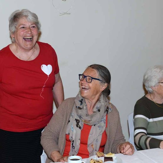 Drei ältere Frauen sitzen lächelnd an einem Tisch. Die Frau auf der linken Seite trägt ein rotes Shirt mit Herzdesign. Die Frau in der Mitte trägt eine Brille und einen Schal. Die Frau auf der rechten Seite trägt ein gestreiftes Shirt. Sie sind von einer weißen Wand umgeben.