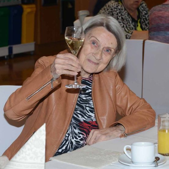 Eine ältere Frau sitzt mit einem Glas Wein am Tisch, lächelt und schaut nach vorne. Sie trägt eine braune Lederjacke. In der Nähe gibt es eine Tasse mit Untertasse, ein Glas Orangensaft und ein gefaltetes Serviettentuch. Andere Menschen sitzen im Hintergrund.