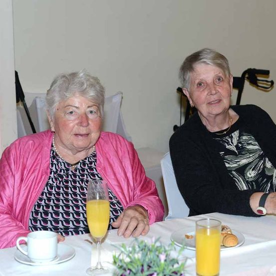 Zwei ältere Frauen sitzen an einem Tisch mit Frühstücksgegenständen. Eine Frau trägt eine rosa Jacke und hat ein Glas Orangensaft. Die andere trägt einen schwarzen Pullover und lächelt.