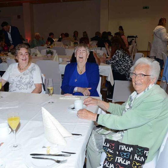 Drei ältere Frauen sitzen an einem Tisch, eine lächelt, eine andere hält ein Weinglas und die dritte hält eine Tasse. Der Tisch ist mit Tellern, Weingläsern und Servietten gedeckt. Andere Personen sitzen im Hintergrund an Tischen.