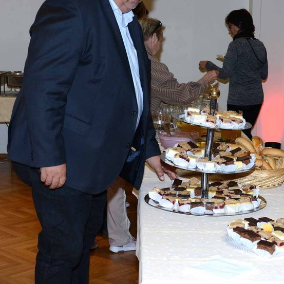 Ein Mann in einem Anzug steht an einem Buffettisch mit einem dreistufigen Kuchenständer. Andere Personen sind im Hintergrund, eine von ihnen greift nach einem Gebäck.