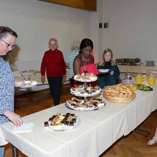 Mehrere Erwachsene und Kinder stehen an einem Buffettisch mit verschiedenen Desserts, Brot und Getränken. Der Raum hat weiße Wände und einen Holzboden.