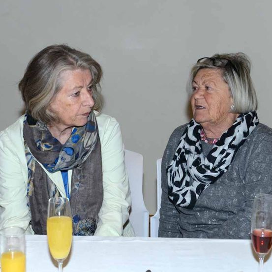Zwei ältere Frauen sitzen an einem Tisch, jede mit einem Glas Orangensaft vor sich. Eine Frau trägt eine gemusterte Halskette. Sie scheinen in ein Gespräch vertieft zu sein.