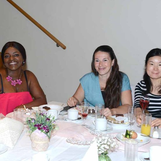 Drei Frauen sitzen an einem Tisch und genießen eine Mahlzeit, mit Blumen und Getränken. Sie lächeln und scheinen eine gute Zeit zu haben. Der Tisch ist mit Tellern, Tassen und Weingläsern gedeckt.