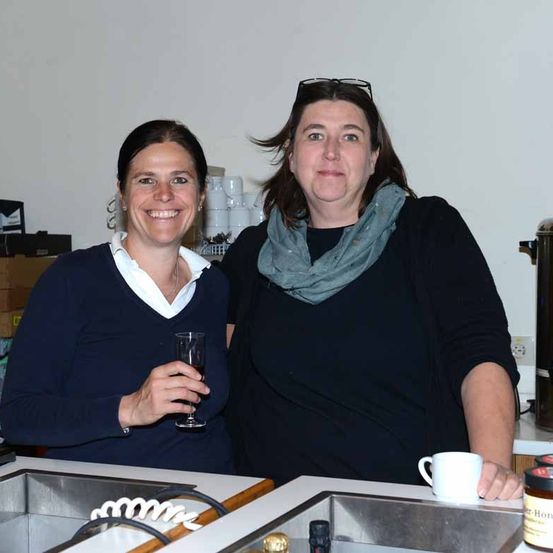 Zwei Frauen stehen lächelnd in einer Küche. Eine hält ein Weinglas, die andere eine Kaffeetasse. Dahinter sind eine Wand und Küchengeräte zu sehen.