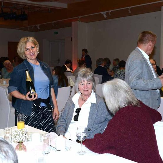 Eine Gruppe von Menschen isst in einem Restaurant. Eine Frau hält eine Flasche Champagner. Eine andere Frau trägt eine Halskette.