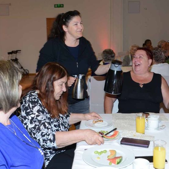 Mehrere Frauen sitzen um einen Tisch und essen. Eine Frau steht hinter ihnen und hält eine Kaffeekanne und ein Glas. Sie alle lächeln.