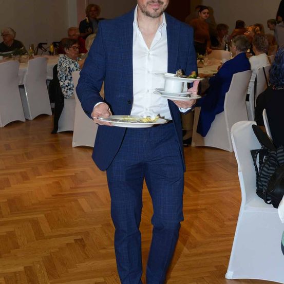 Ein Mann in einem blauen Anzug trägt zwei Teller mit Essen in einem Speisesaal, in dem Menschen auf Stühlen sitzen.