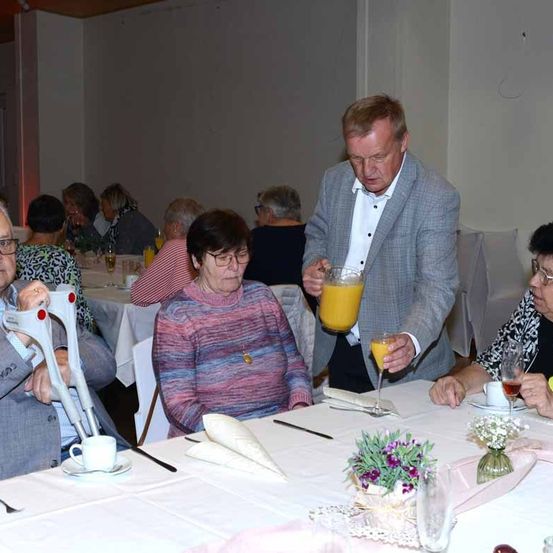 Ein älteres Treffen, ein Mann serviert Getränke an einem Tisch. Eine ältere Frau trägt eine Halskette. Ein Blumenvase steht am Tisch.