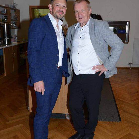 Zwei Männer stehen nebeneinander in einer Küchenecke. Einer trägt einen blauen Anzug, der andere einen grauen Blazer mit weißem Hemd. Sie lächeln und posieren für ein Foto.