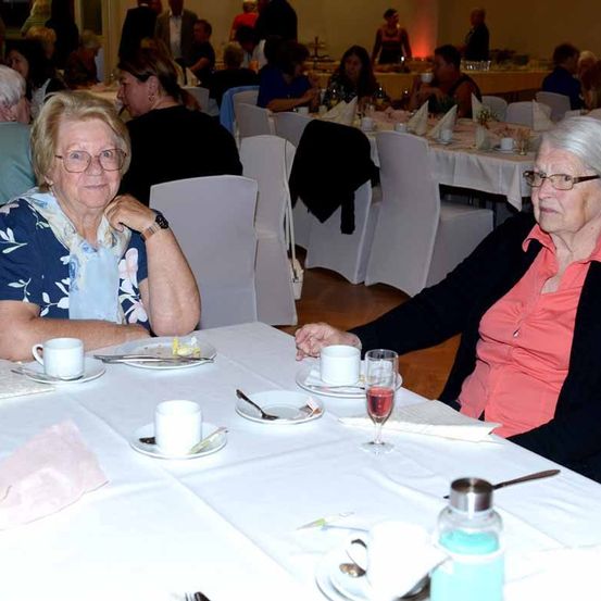 Zwei ältere Frauen sitzen an einem Tisch in einem Esszimmer, lächeln und schauen in die Kamera. Sie sind von anderen Menschen und Tischen mit Tellern und Weingläsern umgeben.