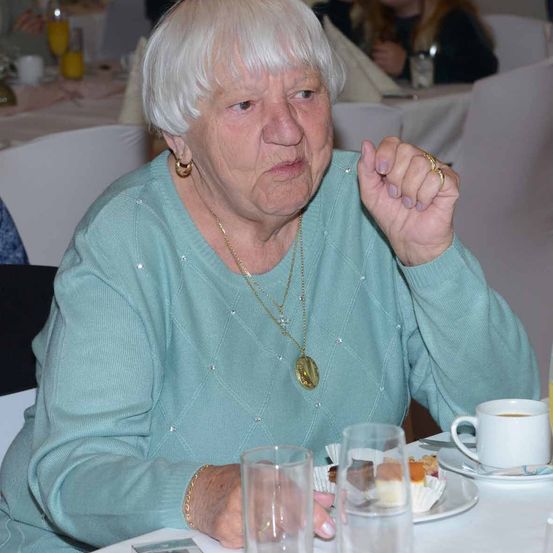 Eine ältere Frau mit weißen Haaren, die einen türkisen Pullover und eine goldene Halskette trägt, sitzt an einem Tisch mit Essen, Getränken und Gläsern. Sie scheint nachdenklich auf etwas zu schauen.