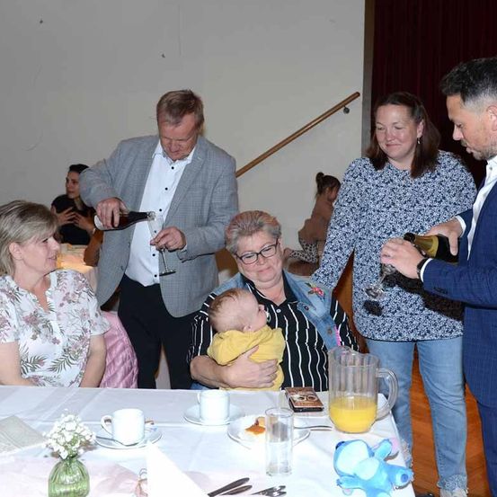 Ein Familientreffen mit Erwachsenen und einem Baby. Ein Mann gießt Wein in ein Glas, während ein anderer eine Champagnerflasche hält. Eine Frau lächelt und hält ein Baby. Der Tisch hat Tassen, Teller und eine Saftschale.