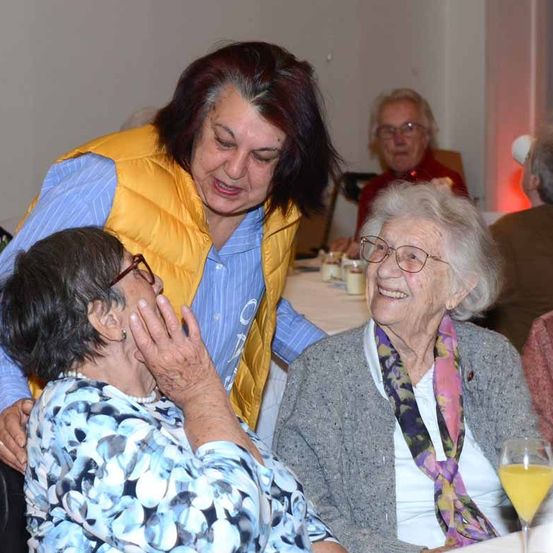 Zwei ältere Frauen sitzen eng beieinander, lächeln und teilen möglicherweise einen Witz. Eine Frau hat ein Glas Orangensaft. Im Hintergrund sitzt eine Frau mit Brille an einem Tisch.