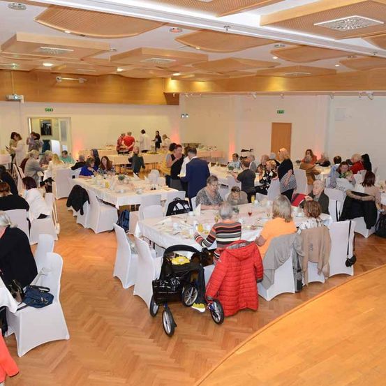 Ein großer Raum mit Holzböden beherbergt ein gesellschaftliches Treffen mit vielen Tischen, an denen mehrere Personen sitzen und sich unterhalten. Einige Personen stehen, während andere auf Stühlen sitzen.