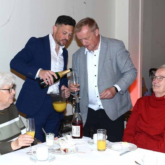 Zwei Männer bereiten sich darauf vor, älteren Menschen, die an einem Tisch sitzen, Champagner einzuschenken, während andere Gläser mit Orangensaft halten.