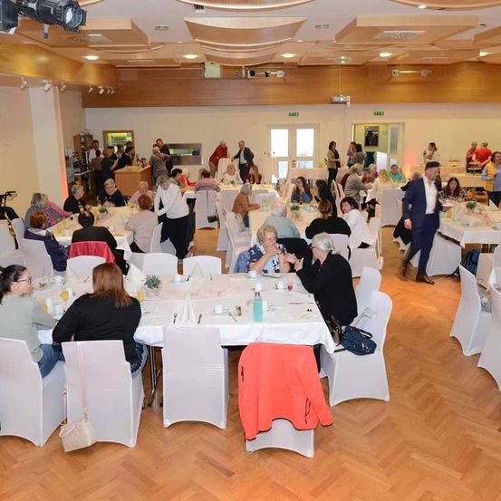 Ein großer Raum mit Holzböden beherbergt eine Versammlung von Menschen, die an Tischen sitzen. Viele unterhalten sich, und ein Mann in Anzug geht vorbei. Tische sind mit weißen Tischdecken und verschiedenen Gegenständen wie Flaschen und Gläsern geschmückt.