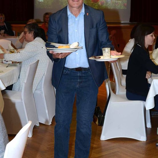 Ein Mann in Anzug und Jeans steht und hält zwei Teller mit Essen. Hinter ihm schaut eine Frau zu. Andere Gäste sitzen an Tischen. Eine Projektionsleinwand zeigt Bilder.