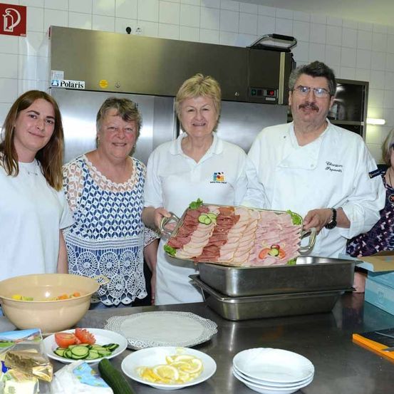 Vier Menschen stehen lächelnd in einer Küche für ein Foto. Sie halten eine Platte mit Fleisch und Gemüse. Verschiedene Teller, Schüsseln und Lebensmittel stehen auf dem Tisch.