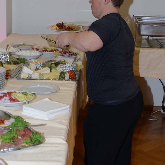 Eine Person in Schwarz serviert Essen auf einem Buffettisch mit verschiedenen Gerichten und Tellern. Der Tisch ist mit einem weißen Tuch bedeckt und hat einen Stapel weißer Servietten.