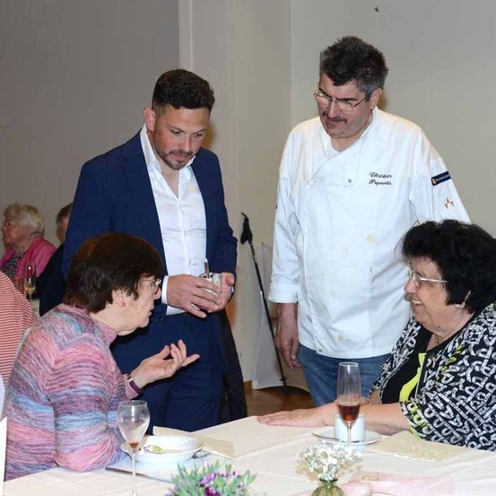 Drei Personen sitzen an einem Tisch mit Weingläsern und einer Schüssel, während ein Koch in einer weißen Jacke hinter ihnen steht. Ein anderer Mann in einem blauen Anzug gestikuliert und spricht mit der Frau in der Brille.