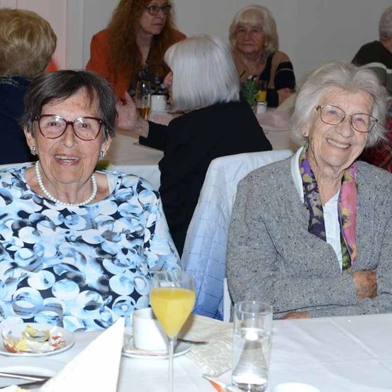 Eine Gruppe älterer Frauen sitzt an einem Tisch, lächelt und sieht glücklich aus. Die Frau vorne trägt eine Brille und eine Perlenkette. Hinter ihr trägt eine andere Frau eine Krawatte. Auf dem Tisch stehen Gläser, Teller und Tassen.